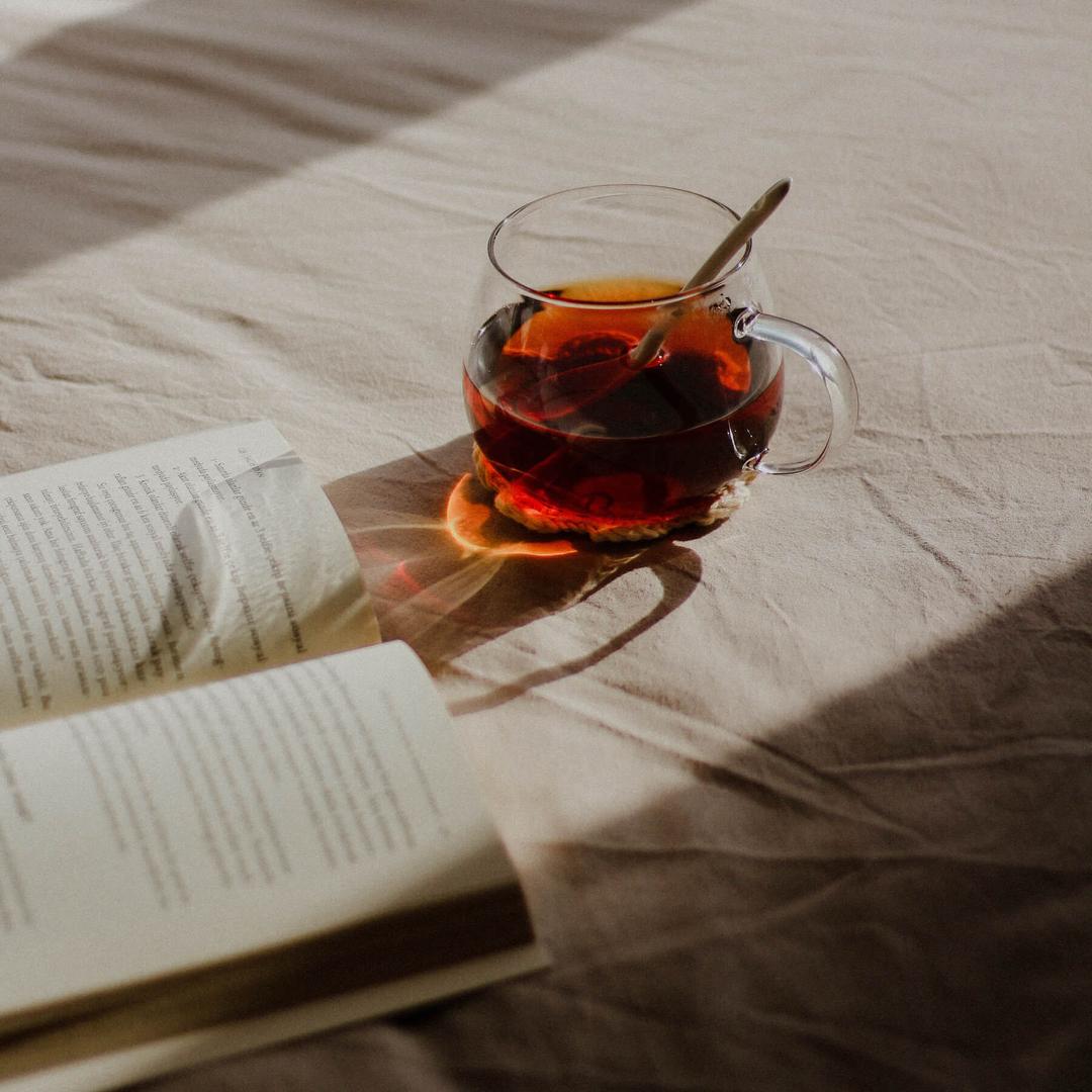 A small glass cup of tea and a book on a white linen bed.